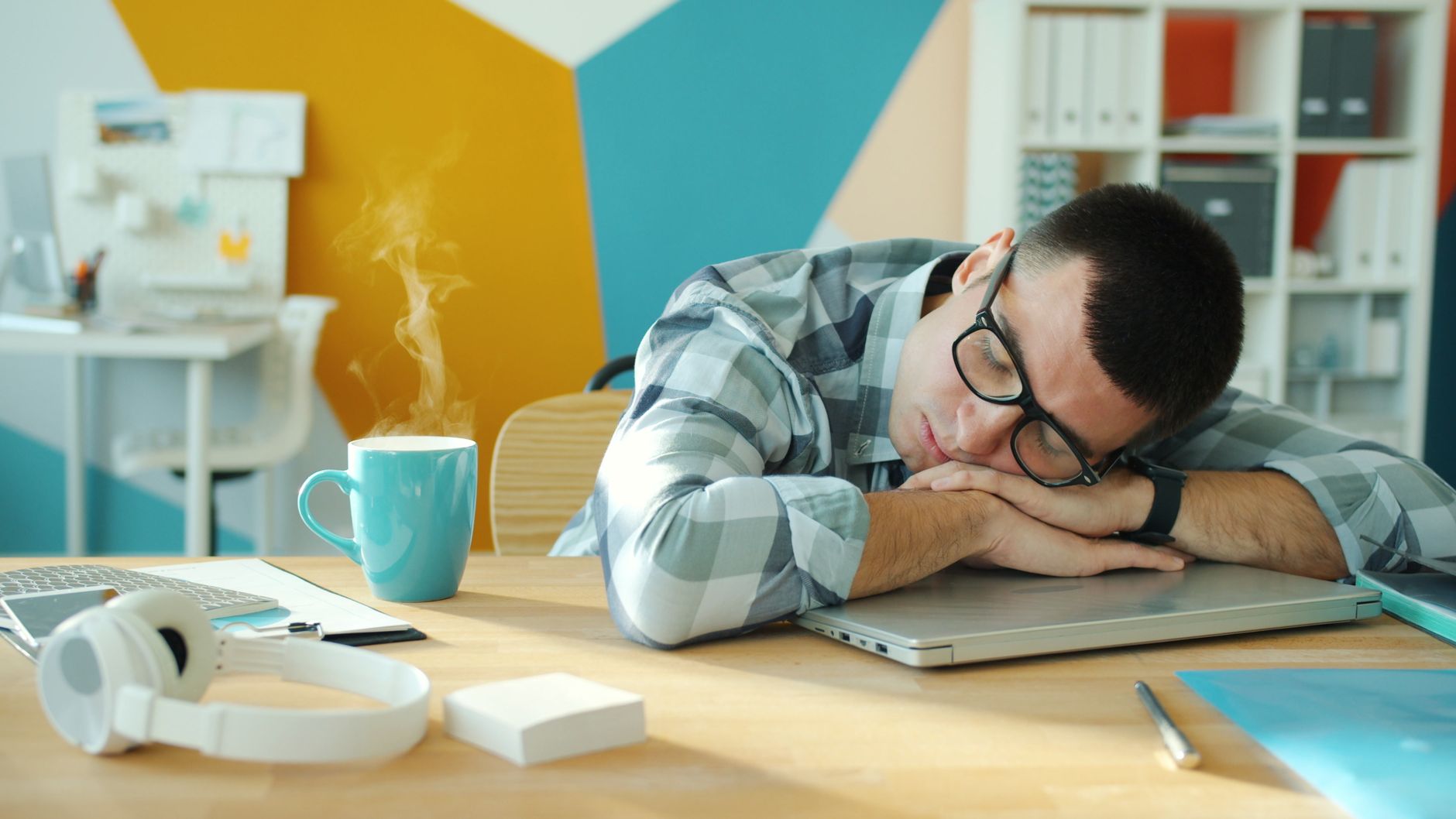 Young man sleeping on a laptop at his desk with a hot beverage nearby.