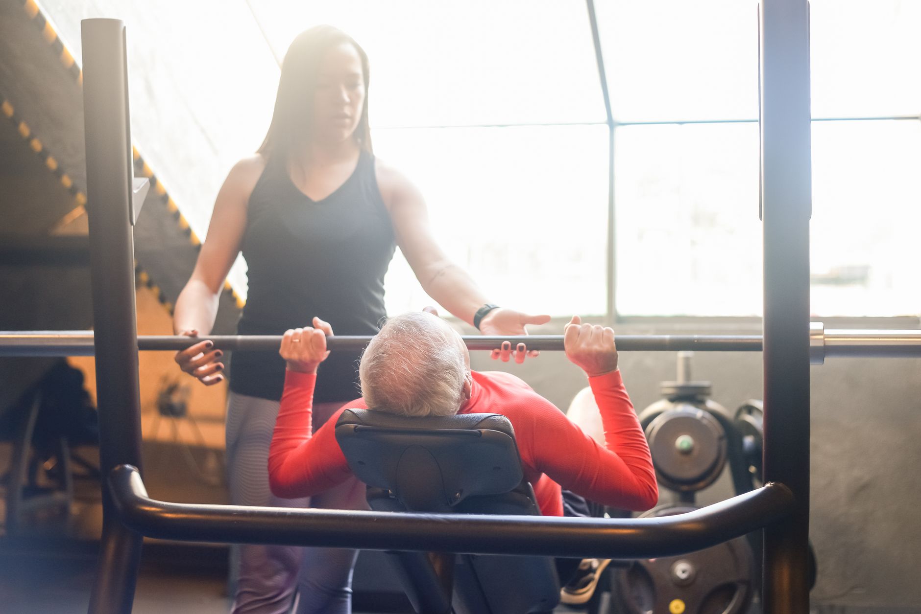 Elderly man in gym performing bench press with female trainer assistance, promoting fitness and health.