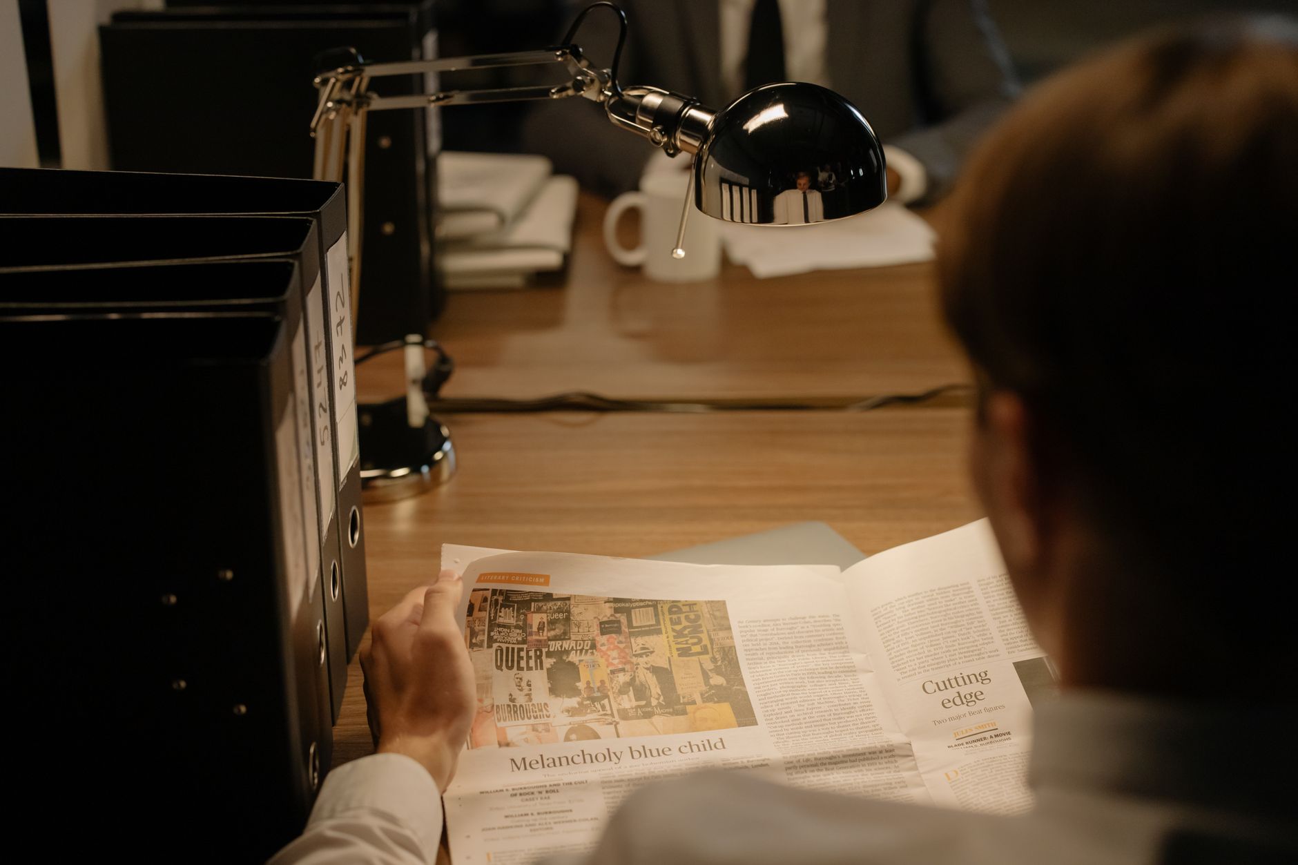 A person reads a newspaper at a desk with a lamp, suggesting investigation work.