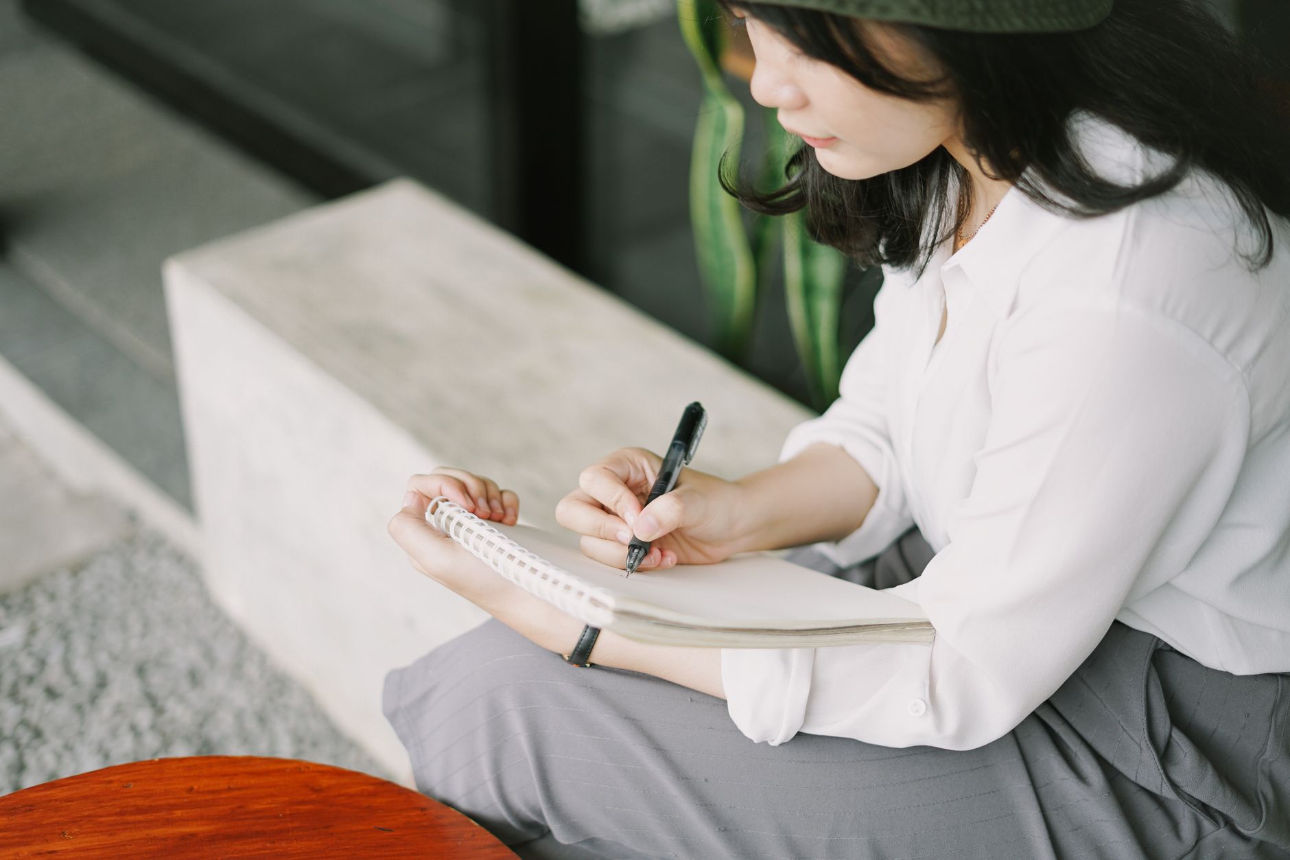 A young woman in a white blouse writing notes in a notebook outdoors.