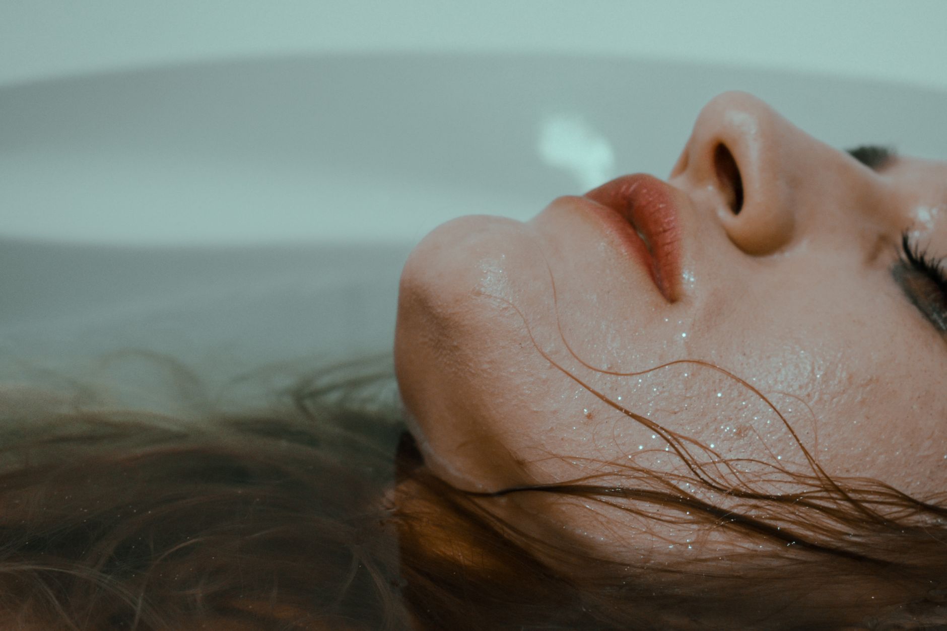 Side view of anonymous lady with wet face and mascara under eyes lying in bathroom with closed eyes with head resting above water