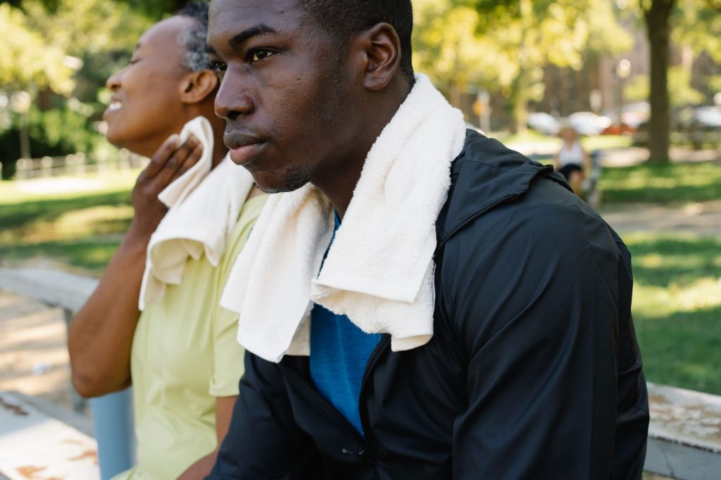 Two adults relaxing on a park bench with towels after a workout under the summer sun.