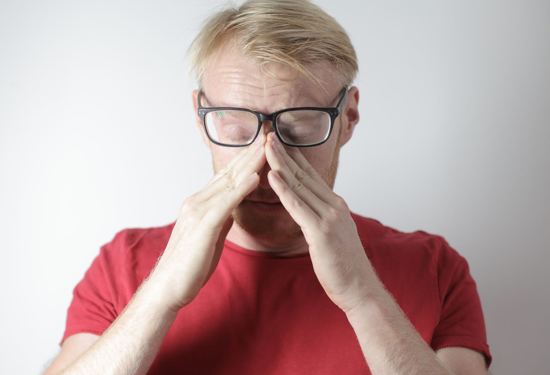 Middle aged male agent with beard in casual clothes and glasses covering nose with hands while spending time at home
