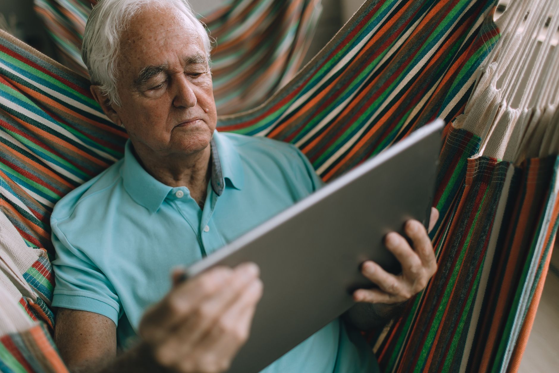 Elderly man enjoys leisure time in hammock while using a tablet, wearing a light blue polo shirt.