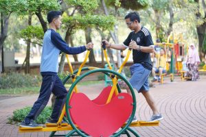 Two adults using outdoor gym equipment in a leafy park setting, enjoying fitness and nature.